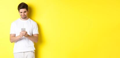 Guy looking displeased at smartphone screen, reading strange message on phone, standing in white t-shirt against yellow background photo
