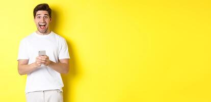 Man looking amazed and happy after reading something on mobile phone, standing over yellow background photo