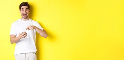 Happy man pointing at smartphone to show promo, check out internet offer, standing over yellow background photo