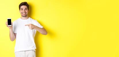 Image of attractive young man pointing finger at smartphone screen, showing an app, standing against yellow background photo