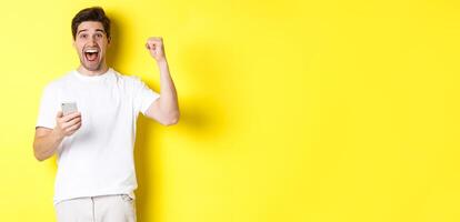Cheerful man winning on smartphone, raising hand up and holding mobile, achieve app goal, standing over yellow background photo