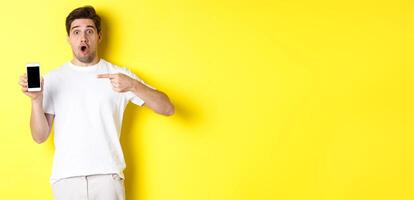 Impressed guy check out smartphone screen, pointing finger at display and gasping amazed, standing over yellow background photo