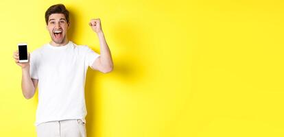 Cheerful guy showing smartphone screen, raising hand up and celebrating, triumphing over internet achievement, standing over yellow background photo