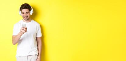 Young man in headphones reading message on smartphone, smiling, listening music in earphones and picking song in app, standing over yellow background photo