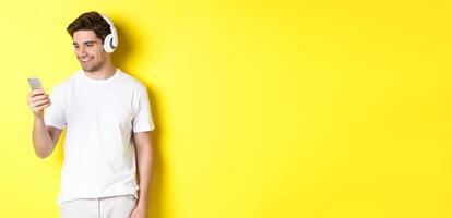 Handsome modern guy choosing playlist on smartphone, wearing headphones, standing over yellow background photo