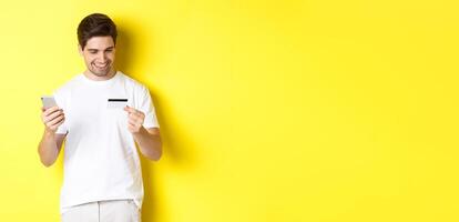 Guy making online order, register credit card in mobile application, holding smartphone and smiling, standing over yellow background photo