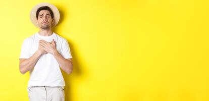 Concept of tourism and summer. Romantic man in straw hat looking nostalgic, close eyes and holding hands on heart, standing against yellow background photo