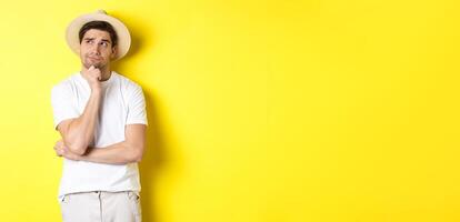 Concept of tourism and summer. Thoughtful man tourist pondering, looking at upper left corner and thinking, standing in straw hat and white t-shirt against yellow background photo