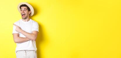 Concept of tourism and lifestyle. Excited handsome guy in straw hat checking out advertisement, pointing and looking at upper left corner logo, yellow background photo