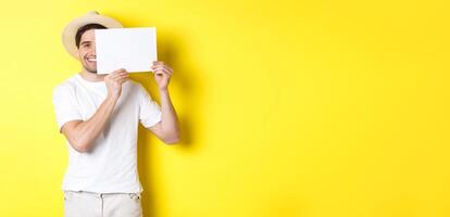 Happy guy on vacation showing blank piece of paper for your logo, holding sign near face and smiling, standing against yellow background photo