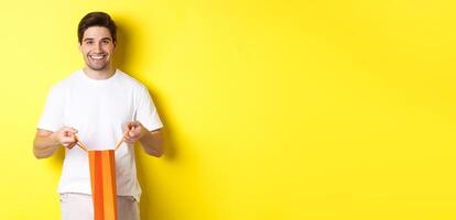 Happy young man open shopping bag with present, smiling at camera, standing against yellow background photo
