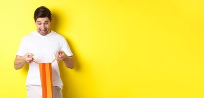 Excited guy open bag with gift, looking inside with amazement and happy face, standing against yellow background photo