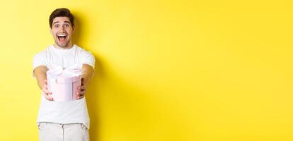 Concept of holidays and celebration. Happy man wishing happy new year, giving you gift, standing over yellow background photo