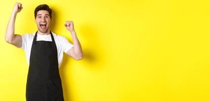 Happy barista celebrating victory, raising hands up and shouting for joy, wearing black apron, shop uniform, standing against yellow background photo