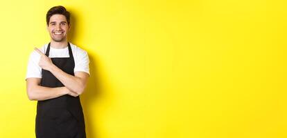 Happy barista pointing finger left and smiling, wearing black apron uniform, standing against yellow background photo