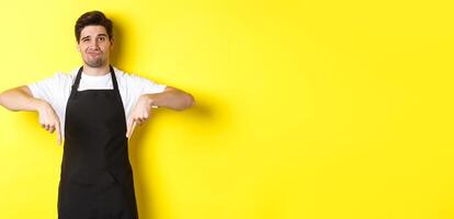 Skeptical barista in black apron pointing fingers down at bad product, looking displeased and unamused, standing over yellow background photo