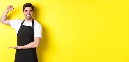 Smiling handsome barista showing something long or large, standing over yellow background photo