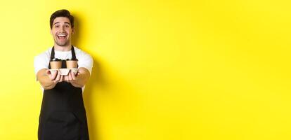 Friendly barista in black apron giving takeaway order, holding two cups of coffee and smiling, standing over yellow background photo