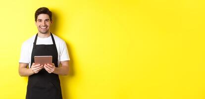 Waiter in black apron taking orders, holding digital tablet and smiling friendly, standing over yellow background photo