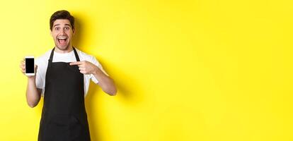 Handsome barista in black apron pointing finger at mobile screen, showing app and smiling, standing over yellow background photo