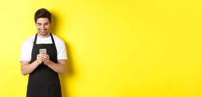 Waiter in black apron reading message on mobile phone, smiling happy, standing over yellow background photo