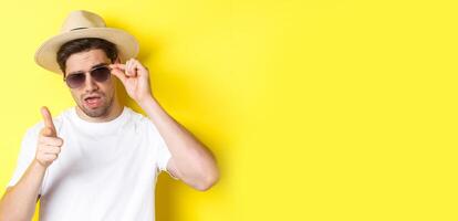Concept of tourism and vacation. Cool and sassy man flirting with you, wearing sunglasses and pointing finger at camera, standing over yellow background photo