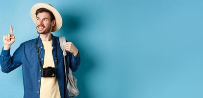 Handsome positive guy on vacation, wearing summer hat, looking and pointing up at logo, smiling pleased, standing with camera and backpack on blue background photo