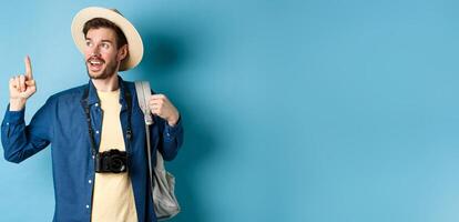 Handsome tourist looking and pointing up, travelling on summer vacation with backpack and camera, standing on blue background photo