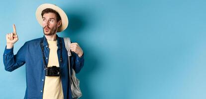 Handsome tourist looking curious and pointing up, showing interesting sightseeing on summer vacation, wearing straw hat and holding backpack with camera, blue background photo