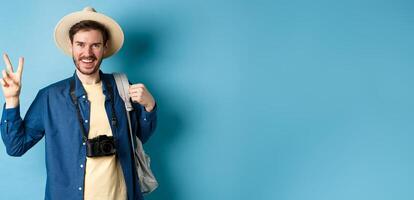 Happy handsome man taking photo on summer vacation, showing peace sign and smiling, wearing straw hat and holding tourist backpack, blue background