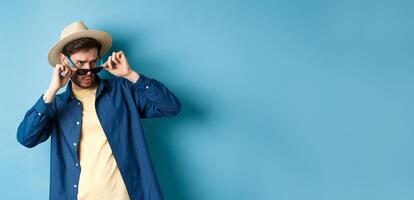 Portrait of guy on vacation staring aside with shocked and cringe face, take-off sunglasses and look at empty space on blue background photo