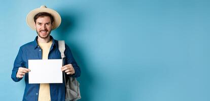Happy tourist with backpack showing empty piece of paper, smiling at camera, standing on blue background. Concept of summer holiday and vacation photo