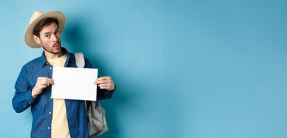 Excited tourist in straw hat, hitchhiking, showing blank piece of paper and looking amused, standing on blue background photo