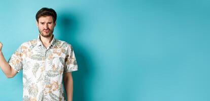 Disappointed tourist cringe and frown, pointing finger aside at empty space, complaining on bad thing, standing in hawaiian shirt on blue background photo