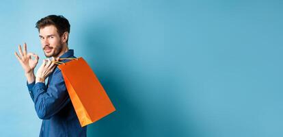 Handsome young man holding shopping bag on shoulder, turn around to look at camera and show okay gesture, recommending shop, blue background photo