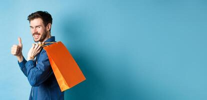 Cheerful shopper holding orange shopping bag on shoulder, turn around at camera with thumbs up, recommending store, blue background photo