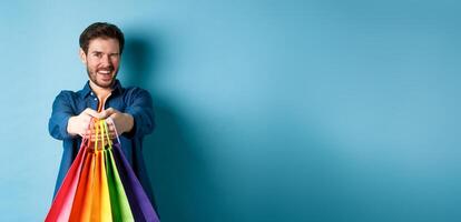 Cheerful handsome man winking and smiling, extending hands with colorful shopping bags, standing on blue background photo