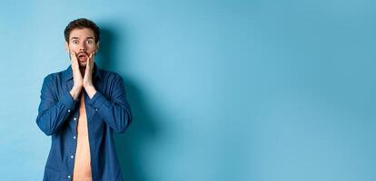 Portrait of guy gasping and dropping jaw with shocked face, staring at camera startled, standing on blue background photo