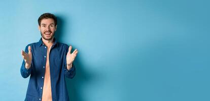 Cheerful young man pointing hands at camera and smiling, feeling glad for you, praising nice work, standing on blue background photo