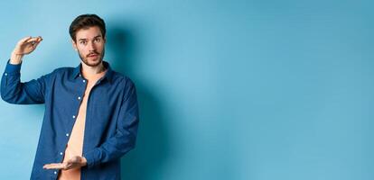 Modern guy showing big size, shaping large object and looking impressed at camera, standing on blue background photo