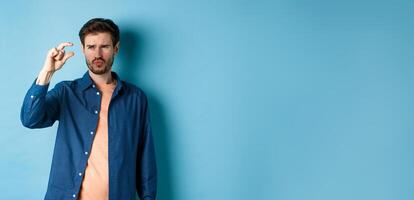Confused guy showing small size and looking doubtful or skeptical, standing over blue background photo