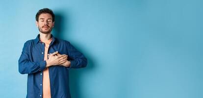 Romantic and nostalgic guy holding hands on heart with eyes closed, keep warm memories, standing on blue background photo
