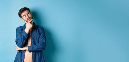 Pensive young man touching face and looking aside at empty space, imaging something, thinking off plan, standing on blue background photo