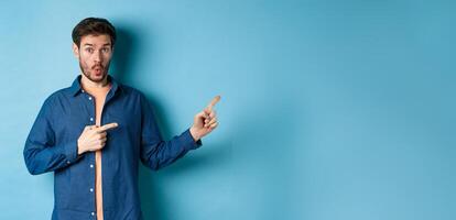 Surprised young man checking out promo, pointing fingers right at logo and saying wow, standing on blue background photo