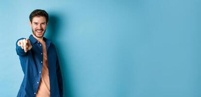 Confident smiling guy choosing or inviting you, pointing finger at camera decisive, standing on blue background photo