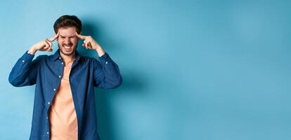 Man feeling sick, touching head and grimacing from painful migraine, having headache or feel dizzy, standing on blue background photo