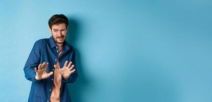 Man cringe and stay away from something nasty, looking aside and stretch out hands to reject bad deal, standing on blue background photo