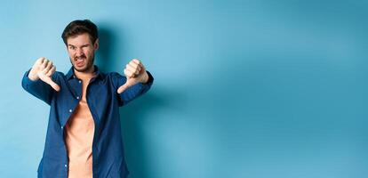 Disgusted guy express negative opinion, showing thumbs down and tongue, frowning upset, disapprove something bad, standing on blue background photo