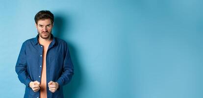 Angry and pissed-off man clench fists and stare with disdain at camera, trying to keep himself together, look with rage, standing on blue background photo