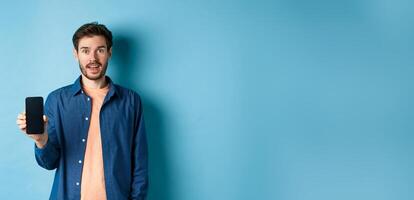 Happy caucasian guy showing empty mobile screen and smiling, standing on blue background photo